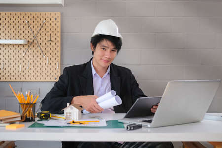 Handsome male architect in formal suit and hardhat with rolled blueprints sitting at workplace and smiling to camera.の写真素材