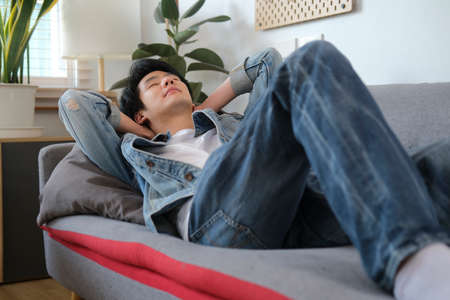 Pleased young asian man with hands behind head relaxing on couch at home.の写真素材