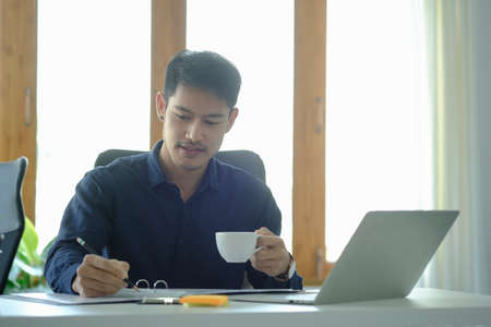 Successful young businessman drinking coffee and checking financial reports at office desk.の写真素材