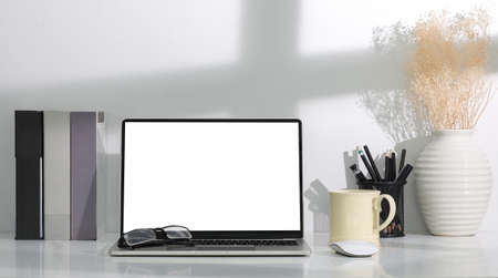 Modern workplace with computer laptop, books, coffee cup and pencil holder on white table.
