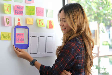 Focused female web developer is planning application for smart phone on whiteboard at creative...