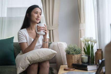 Positive young woman sitting on comfortable couch at home and drinking hot coffee in the morning.の写真素材