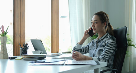 Smiling female entrepreneur sitting at bright modern office and talking on mobile phone.の写真素材