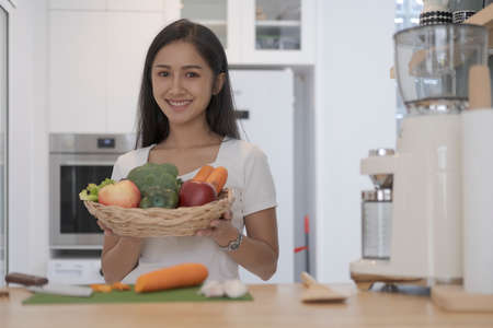 Smiling young woman holding basket with fresh vegetables standing at kitchen counter. Healthy...
