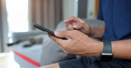 Cropped shot man in casual clothes sitting on sofa and using smart phone, checking social media,...