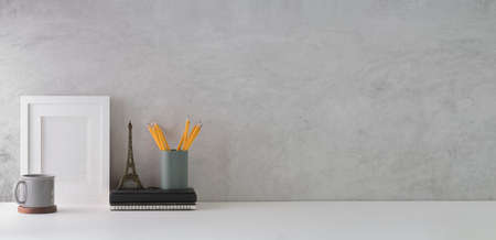 Stylish workplace with blank picture frame, pencil holder, book and coffee cup on white table against loft wall. Copy space for your text.の写真素材