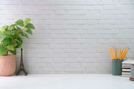 Minimal workplace with potted plant, pencil holder and books on white table against brick wall. Copy...