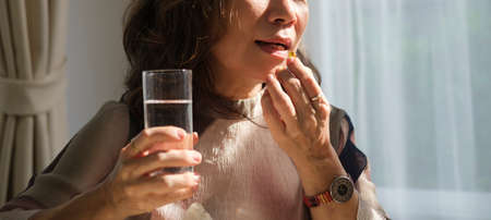 Mature woman woman holding pill and glass of water, taking daily vitamins or dietary supplements. Elderly healthcare, medication concept.の写真素材