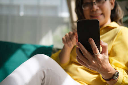 Smiling middle aged mature woman enjoying using mobile apps, browsing social media while sitting on sofa at home.の写真素材