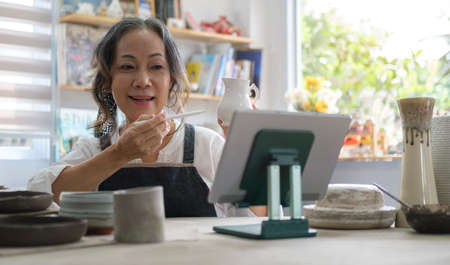 Mature woman holding pottery earthenware product in front of digital tablet while live streaming....