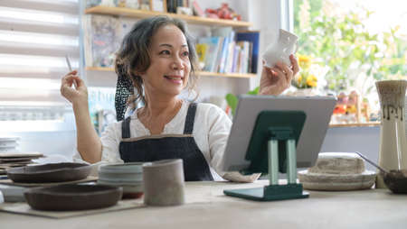 Smiling senior female holding pottery earthenware product in front of digital tablet while live...