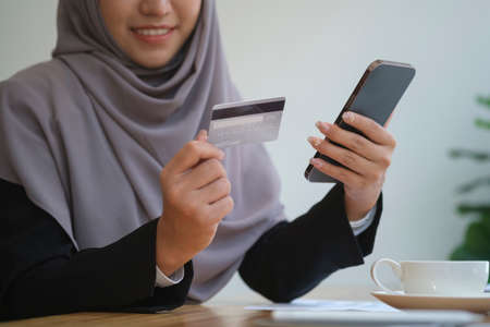 Smiling muslim businesswoman in hijab holding credit card for online shopping on her smart phone....
