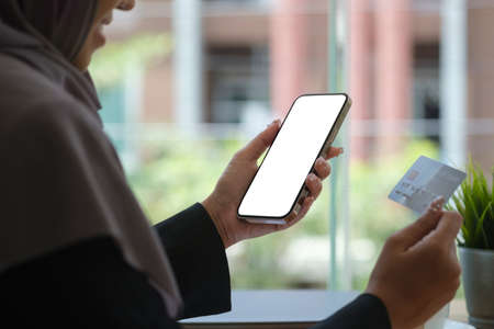 Cropped shot young muslim woman in hijab holding credit card and smart phone with blank screen....
