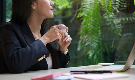 Business woman drinking hot coffee sitting in modern workplace and looking out the window, enjoys view, thinking work plan.の写真素材