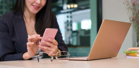 Smiling businesswoman sitting at her office desk front of laptop computer and using smart phone.