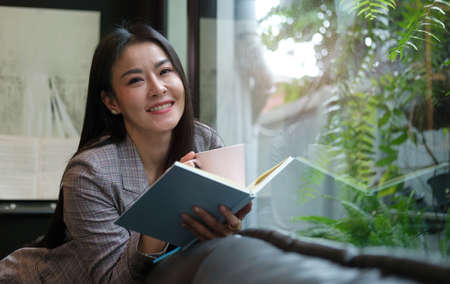 Happy young woman relaxing on sofa in front of window reading book and drinking coffee.
