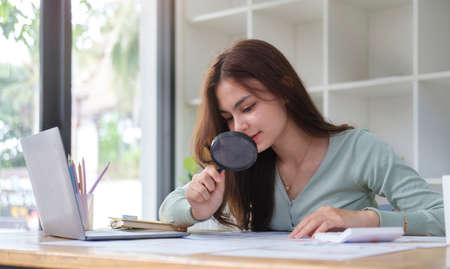 Young female investor examining financial documents with magnifying glass.の写真素材