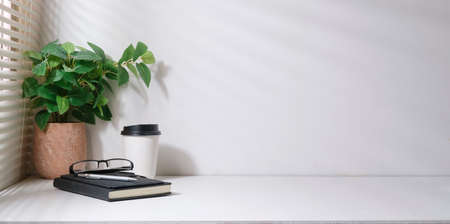 Minimal workplace, houseplant, notebook, glasses and coffee cup on white table with sunlight through blinds by the window.の写真素材