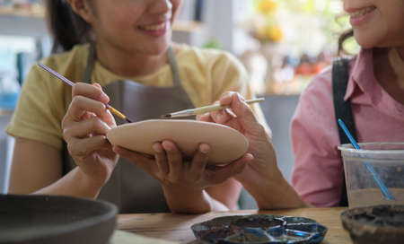Smiling young female ceramist drawing on ceramic bowl in pottery workshop. Hobbies and indoors leisure activity concept.の写真素材