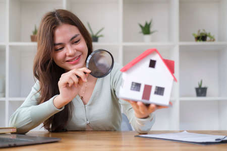 Smiling young woman looking at house model with magnifying glass. Real estate appraisal, land valuation and house selection concept.の写真素材