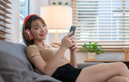 Cheerful young woman wearing headphones and using smart phone in cozy living room.の写真素材