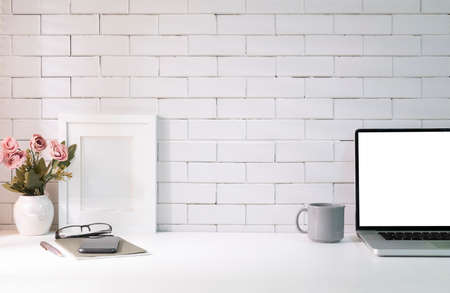 Laptop with blank screen, picture frame, flower pot and notebook on white table. Feminine workplace.