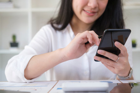 Smiling female employee sitting in bright office and using smart phone for communicating on social network.の写真素材