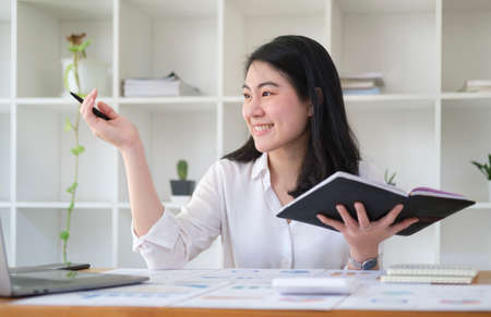 Smiling millennial businesswoman holding notebook and looking out the window, enjoys view or...