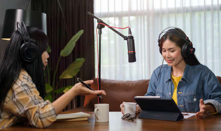 Two young women radio hosts using microphone and audio mixer recording podcast at home studio...