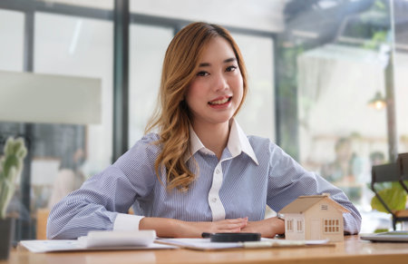 Smiling young female investor sitting at her workplace with house model and calculator. Real estate and property concept.の写真素材