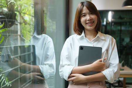 Positive young businesswoman holding digital tablet and smiling at camera while standing in modern...