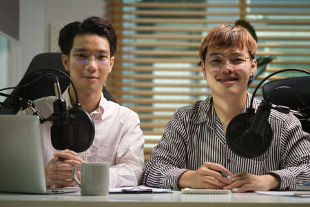 Two radio hosts sitting front of microphone and laptop at their broadcasting studio.