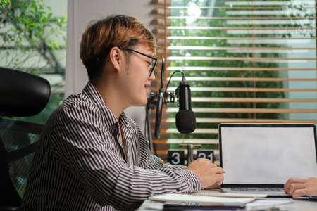 Young man vlogger using microphone and laptop streaming live audio podcast in home studio.