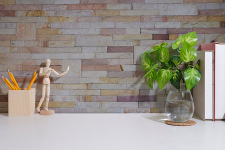 Home office desk. Books, pencil holder, glass of water and houseplant on white table with brick...