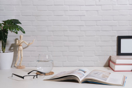 Open book, eyeglasses, glass of water and houseplant on white table with brick wall.