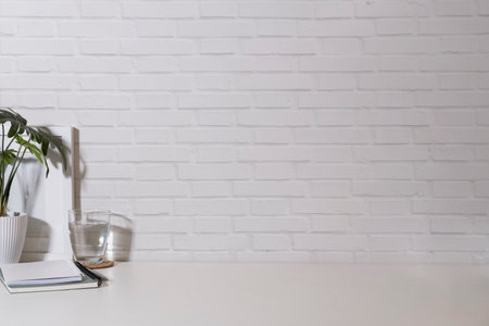 White picture frame, potted plant, glass of water and notebook on white table against brick wall....