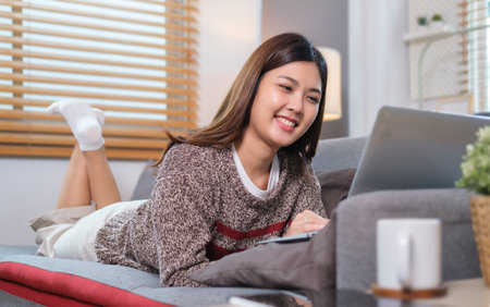 Cheerful young woman lying on couch and surfing internet with laptop.の写真素材