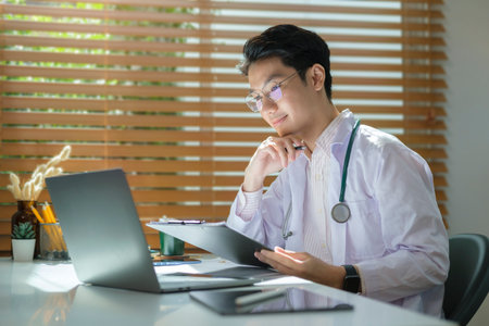 Smiling man doctor wearing white uniform reading electronic medical records on laptop computer.の写真素材