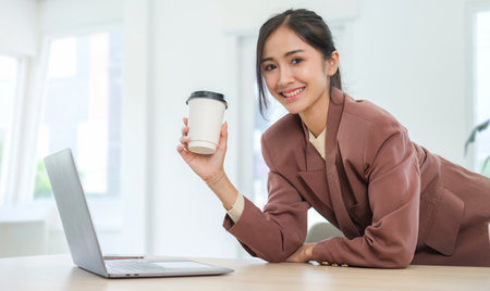 Confident female manager hand holding paper cup, standing front of laptop computer and smiling to camera.の写真素材