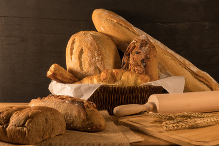 Various crusty bread in wicker basket, wheat  and wooden rolling pin on rustic table.