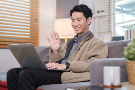 Happy man waving hand, having video call on laptop while sitting on couch in cozy living room.の写真素材