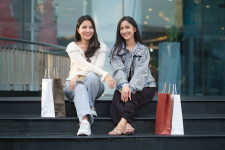 Two young women in trendy casual clothes sitting on stair in front of the shopping mall and looking...