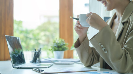 Smiling millennial businesswoman drinking and enjoying coffee during working on laptop in bright modern office.の写真素材