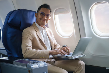 Smiling businessman in elegant suit using laptop while sitting in airplane cabin.