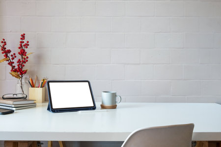 Minimal workspace with digital tablet, books, coffee cup and stationery on white desk against brick...