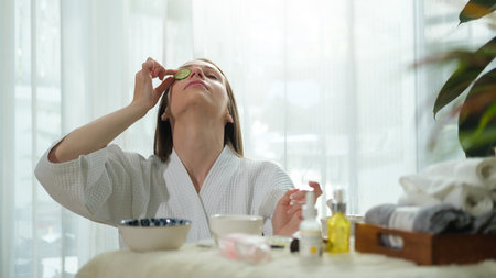 Young woman in bathrobe putting fresh cucumber on her eye while doing beauty treatment at home.