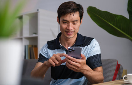 Happy millennial asian man typing message or ordering food delivery on smart phone while sitting in cozy home.の写真素材
