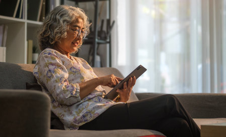 Beautiful mature woman sitting in bright living room and using digital tablet, surfing internet, checking news in the morning.の写真素材