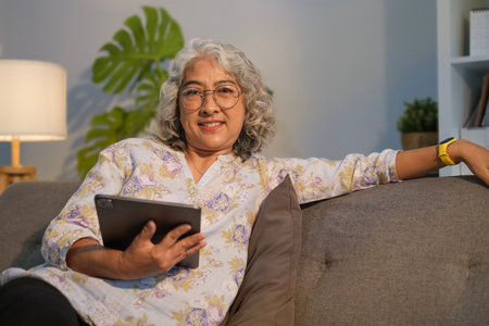 Satisfied mature woman surfing internet on digital tablet while relaxinf on couch at home.の写真素材