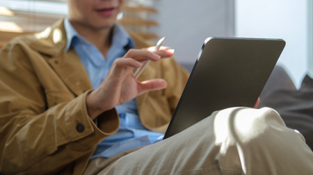 Cropped shot young man sitting in bright living room and using digital tablet.の写真素材
